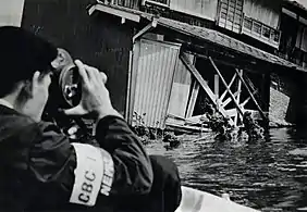 Black and white image of a cameraman in flood waters taking an image of a building's damaged façade.