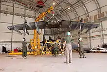 Yellow crane hoisting a wing of an aircraft, with several people standing below securing the wing. This takes place inside an aircraft hangar.