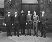 Six men in suits and ties standing in front of a building