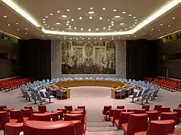 A meeting room, in the centre is a round table with blue chairs. In the background are red chairs.