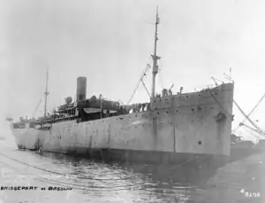 A photograph of a ship tied up at a pier. The bow of the ship is on the right side of the frame with mooring lines running to the dock. On the left of the frame is the calm water of a harbor.