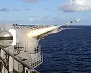 Firing of a missile from Theodore Roosevelt while at sea, seen from the flight deck. There is a Phalanx cannon on the left, with a white, domed upper section and a black cannon on the lower part.