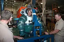 A diver wearing the Oceanworks ADS 2000 suit with the helmet dome open stands in an indoor test pool and talks to two other naval officers