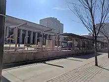 A train sits in a modern railway station in front of a stately stone railway depot on a clear day.