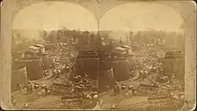 Two identical photos of a street, crowded with various horse-drawn wagons, with large storage tanks on either side and oil derricks visible in the distance. The images are mounted side-by-side on a card.