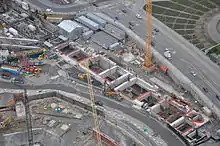 A construction site with several cranes and walls surrounding a long, rectangular hole in the ground.