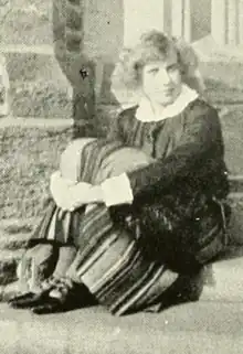 A young white woman with fair hair, seated outdoors in front of a stone building, wearing a dark top and striped skirt