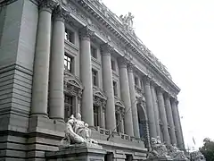 An oblique view of a gray stone neoclassical building. There are 12 columns across the front of the building spaced equally except for the corners and on either side of the central door where they are doubled up.