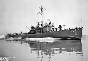 USS PC-815, a single-masted vessel running at speed with a large wake, and with a deck gun prominent on the bow