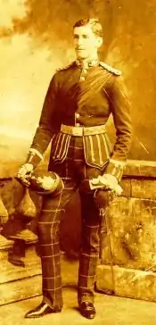 Formal studio sepia portrait photograph of Gustavus Hamilton Blenkinsopp Coulson in military dress uniform with checked trousers, pointed black shoes, helmet in hand, other hand on hilt of sheathed sword, standing in front of stone balustrade