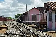 Imagem de um trilho de trem em primeiro plano e, à direita, edificação antiga de parede rosa com an estação e an inscrição Bebedouro em branco