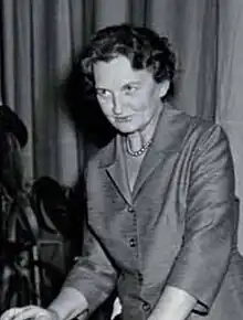 A short haired woman in a suit, wearing pearls, leaning on a podium.