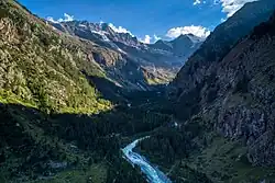 A remote hut is made of basic wood with a slanted roof. It is surrounded by coniferous trees and mountains. A winding path leads from the foreground to the background where the mountain Gran Paradiso is seen.