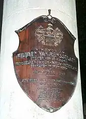 A white, cylindrical stone is marked with a copper-colored shield marking the burial place of Van Rensselaer, which includes Dutch text.