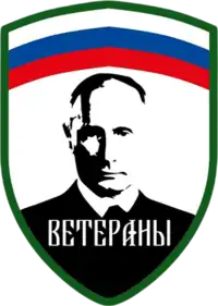 The Logo and patch of the Redut formation "Veterany", with the Russian tricolour at the top horizontally, the edges being green and the below part with white background and a black portrait of Putin in the middle, with the text "Veterany" or "Veterans" written below him.