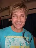 A man in a light blue t-shirt smiles towards the camera. He has blond tousled hair, and sports a stubble.