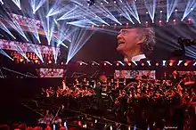 Large concert hall with an orchestra playing, and the conductor's face projected on a large screen over the stage