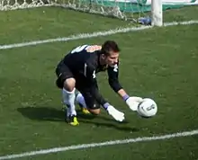 Man with light skin and short black hair, wearing a goalkeeper football kit composed of black shirt and shorts and white socks, crouching in front of a goal with his gloved hands stretched towards a moving football.