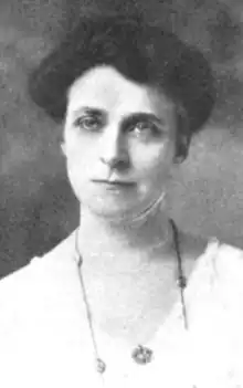 A white woman with dark hair in an updo, wearing a v-neck white garment and a necklace