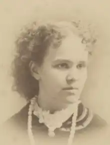 A young white woman with curly hair parted center. She is wearing a high frilly white collar and a strand of large, light-colored beads.