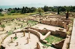 The white stone remains of multiple buildings set in a grassy area surrounded by trees with the sea in the background