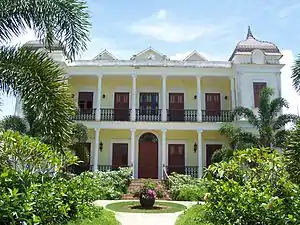 Photograph of the Hacienda Iruena Manor House, a large, two-story house with verandas on both levels and a corner tower, set in a lushly vegetated setting