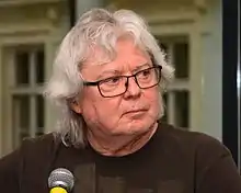 white-haired, older white male wearing glasses, sitting behind microphone, looking left of camera