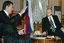 Two men in suits sit talking around a low wooden table.