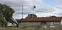 A tan, shed-like building with a red Fox 25 W L A X - T V sign and a small tower with antennas adjacent to it, as seen from a nearby highway