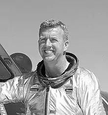 A smiling and energetic blonde man wearing a pressure suit stands in front of an airplane