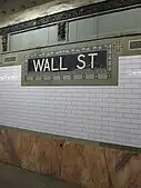 View of the wall along the platform. There is a a pink marble wainscoting on the lowest part of the wall, with white glass tiles above. A white-on-blue tile plaque with the words "Wall Street" and floral motifs is also placed on the wall.