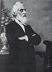 a man with a white beard and hair faces right, with his arms folded. He is dressed in a suit of a sort worn in the late 19th century.