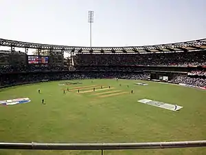 View from the top of a cricket ground during a cricket match; players are visible in the field.