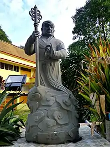 Monk in robes, stern face, bushy eyebrows, holding staff and prayer beads