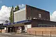 A red-bricked building with a rectangular, dark blue sign reading "WEST ACTON STATION" in white letters all under a blue sky with white clouds