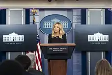 A 32-year-old woman conducting a press conference in the White House, specifically in the James S. Brady Press Briefing Room in  West Wing of the White House.