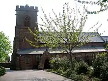 A Gothic Revival sandstone church seen from the side with a crenellated tower on the left and the body of the church with a clerestory to the right; in the foreground on the right is a path with shrubs and a tree that partly obstruct the view of the church