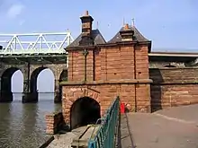 A red sandstone building seen from the rear, in two storeys, with an arch over a footpath on the left in the lower storey, and a plain wall in the upper storey; two chimneys and two hipped roofs; on the left are parts of the bridges and on right is the edge of the approach to the road bridge