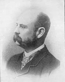 A black-and-white photograph of the head and shoulders of a middle-aged balding man with a large moustache.