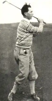 A white man with dark hair, in a golf swing pose, wearing a knitted sweater and short trousers