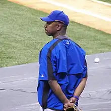 An African-American man, wearing a baseball hat and jacket, stands outside a baseball diamond.