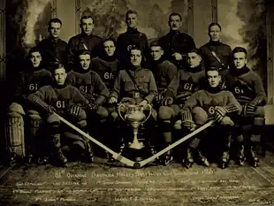 Black and white photo of hockey team sitting behind the Allan Cup trophy