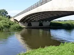 View from grassy riverbank looking up at the natural white concrete arched form of bridge against a pale blue sky on a sunny day