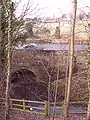 The Wom Brook at Rushford Bridge, where it enters the village under the old Stafford-Worcester Road. St Benedict Biscop's church and the village centre are visible in the background.