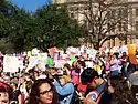 Hundreds of people in light clothes, many holding homemade signs, stand in front of several trees and a light stone building.