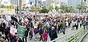 Marchers with signs walk down a street from right to left. Buildings and palm trees stand in the background.