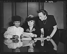 Three women posed for a photo; two seated, one standing and bent over the other two. The table in front of them is reflective; the middle woman is holding papers.