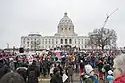 People with flags and signs mill about in front of a statehouse on a butty day.