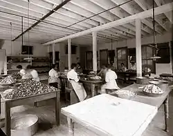 A black-and-white photograph taken from the corner of a room with tables on which small objects are piled, some in containers and others loose. In the middle of the room women in white aprons are seated around the tables, apparently at work.