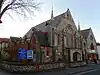 Front view of a wide three-part flint church with red brick dressings. The main (left) section is flanked by stone spires and has a large four-light lancet window with quatrefoils and a sexfoil, and two single-light lancets.  In the middle is a narrow, low entrance section.  To the right is a plain-walled section with three lancets and an arched window.